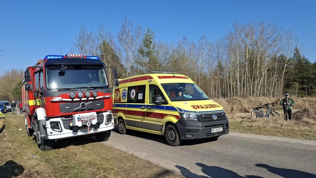 Czerwony wóz strażacki i żółta karetka stoją na drodze, obok grupy osób oraz roweru w tle, w otoczeniu drzew.