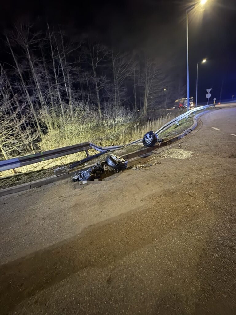 Zniszczony fragment bariery ochronnej oraz odłączone koło leżące na poboczu drogi w nocy, w otoczeniu drzew.