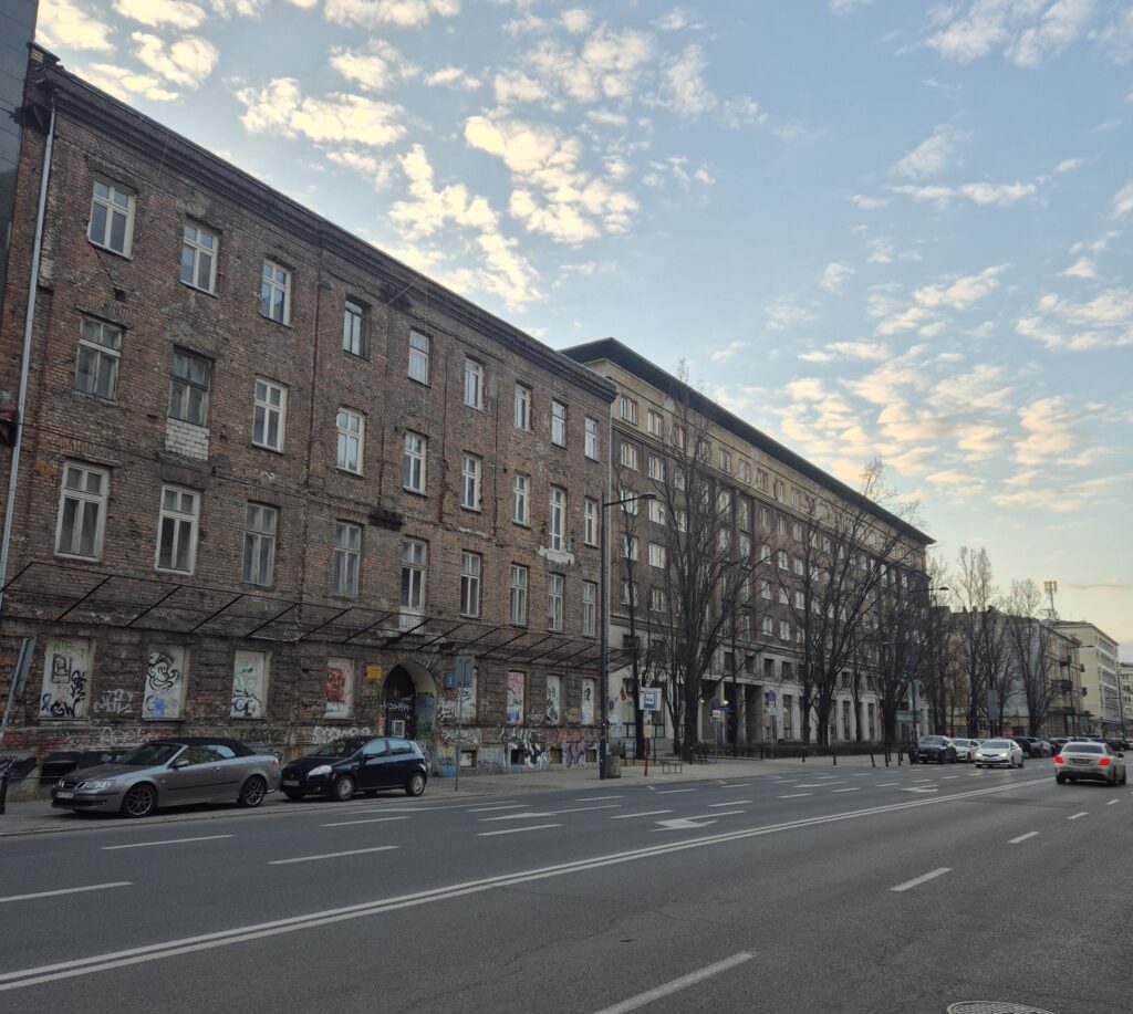 Ceglana kamienica z graffiti na parterze, obok nowoczesny budynek, w tle niebo z chmurami. Ulica z zaparkowanymi samochodami.