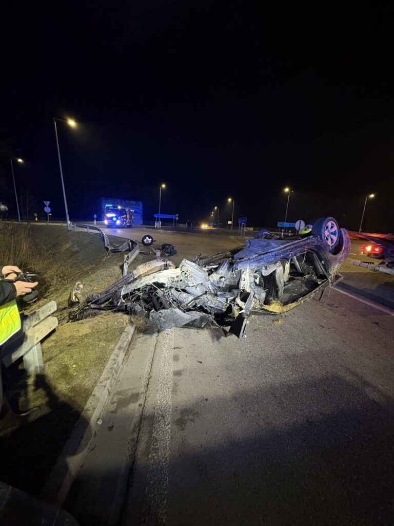 Na drodze leży przewrócone auto osobowe, a w tle widoczne są policyjne światła i zabezpieczenia.