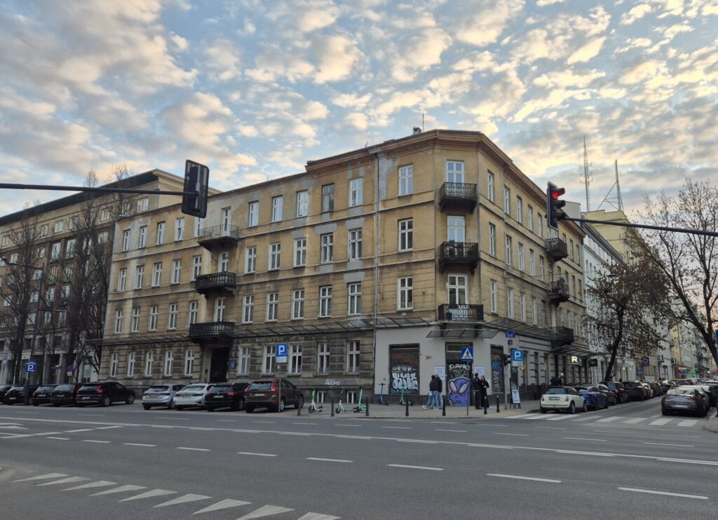 Budynek mieszkalny z balkonami i graffiti na parterze, w tle ruchliwa ulica z zaparkowanymi samochodami.