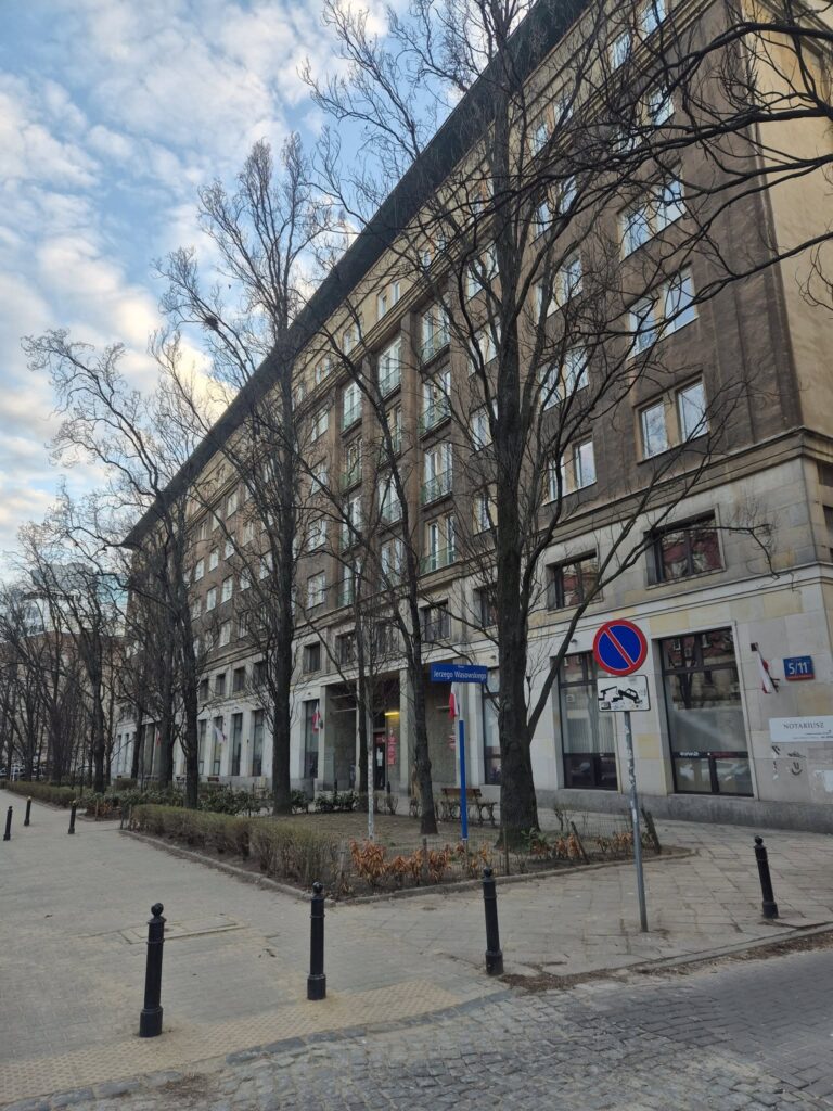 Nowoczesny biurowiec z przeszkloną fasadą otoczony nagimi drzewami w miejskim otoczeniu, z widocznymi znakami drogowymi.