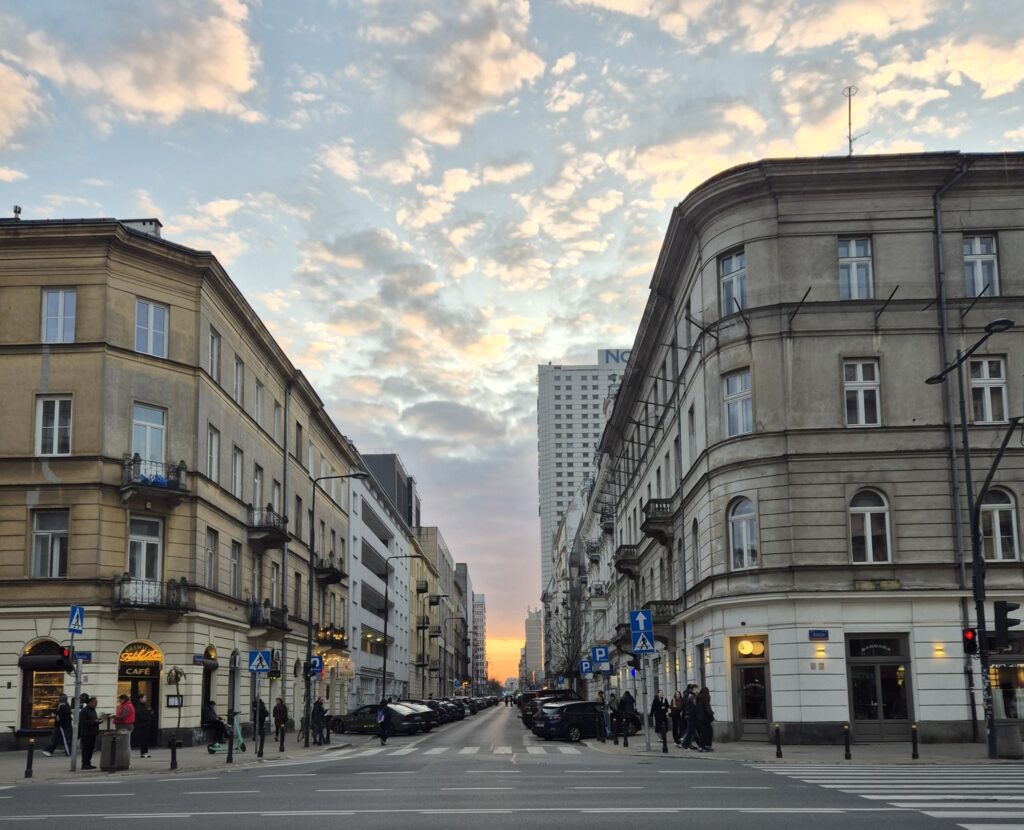 Ulica z zabytkowymi budynkami i nowoczesnymi wieżowcami, w tle zachód słońca z chmurami na niebie.