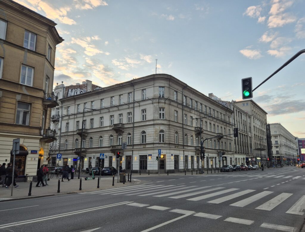 Ulica z nowoczesnym oświetleniem i znakami drogowymi, obok chodnika przechodnie oraz zaparkowane pojazdy.