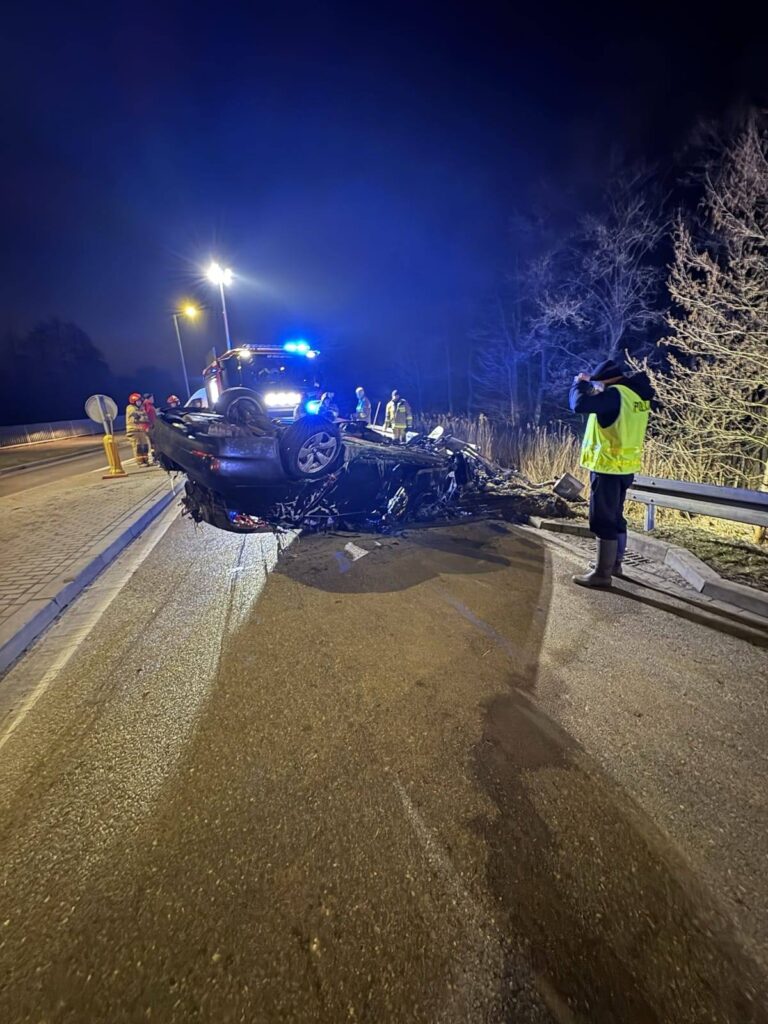 W nocy służby ratunkowe pracują przy przewróconym samochodzie osobowym na poboczu drogi.