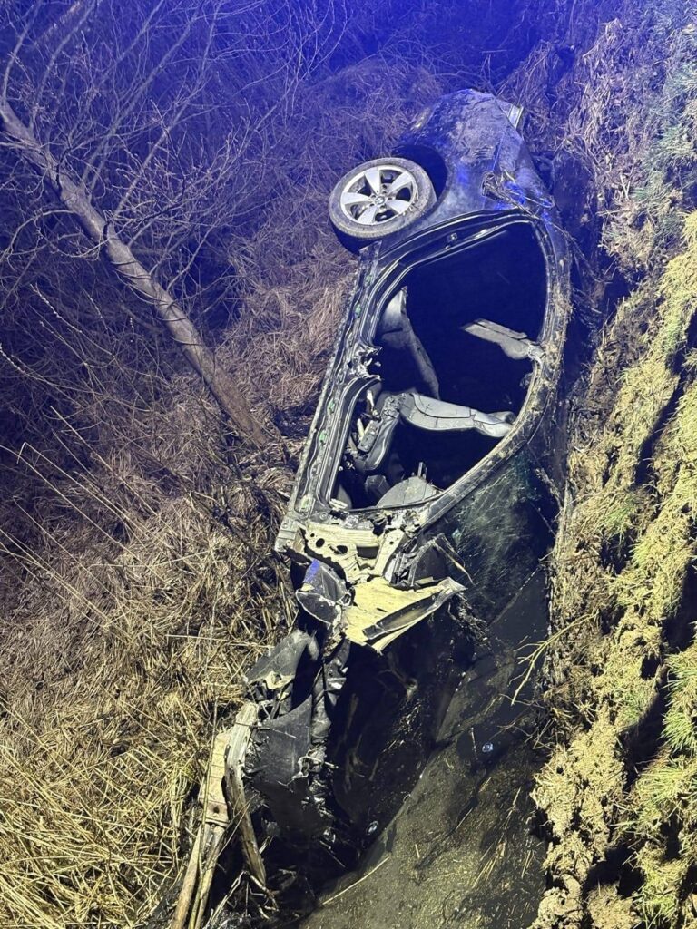 Wrak samochodu leżący na boku w rowie, otoczony przez zarośla i drzewa, z uszkodzoną karoserią.
