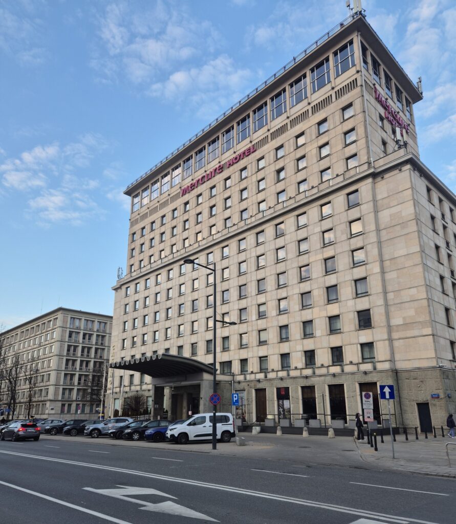 Nowoczesny hotel z przeszkloną fasadą i zadaszonym wejściem, otoczony ulicą i zaparkowanymi samochodami.