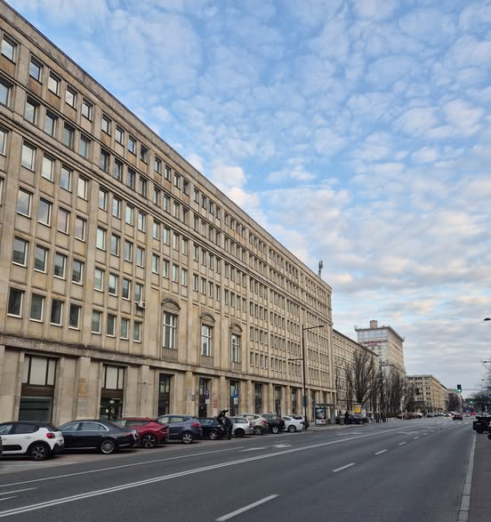 Nowoczesny budynek biurowy z dużymi oknami, widoczny przy ruchliwej ulicy, pod błękitnym niebem z chmurami.