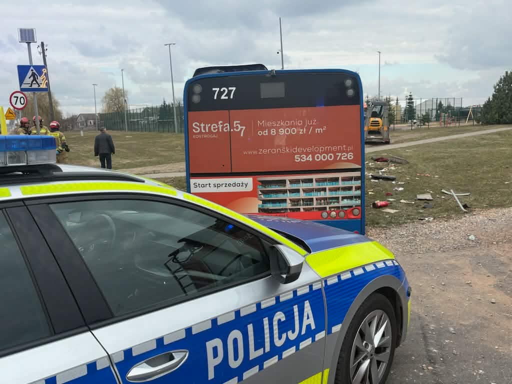 Policyjny radiowóz z włączonymi sygnałami świetlnymi stoi na poboczu, obok przystanku autobusowego z reklamą mieszkań.