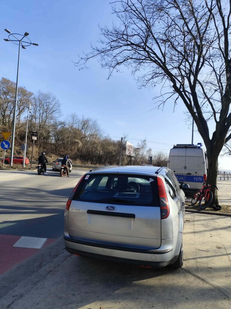 Srebrny samochód osobowy zaparkowany na chodniku obok policyjnego furgonu, w tle widać motocyklistów na drodze.