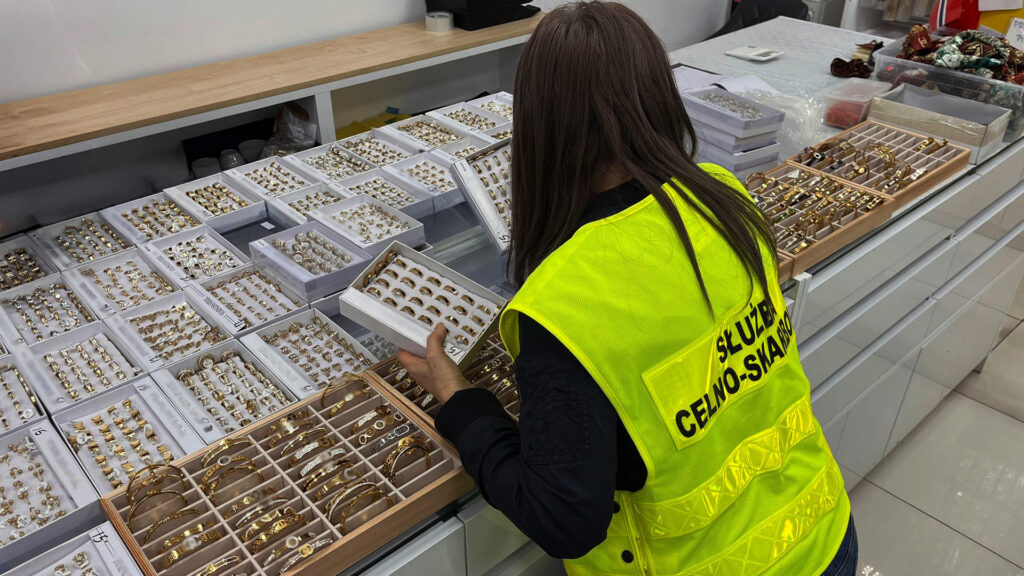 Kobieta w żółtej kamizelce przeszukuje biżuterię w sklepie, analizując zawartość pudełek z ozdobami.