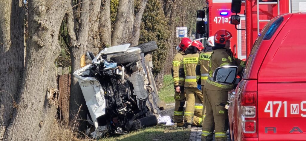 Samochód osobowy przewrócony na bok, obok grupka strażaków w mundurach i kaskach, w tle wóz strażacki.