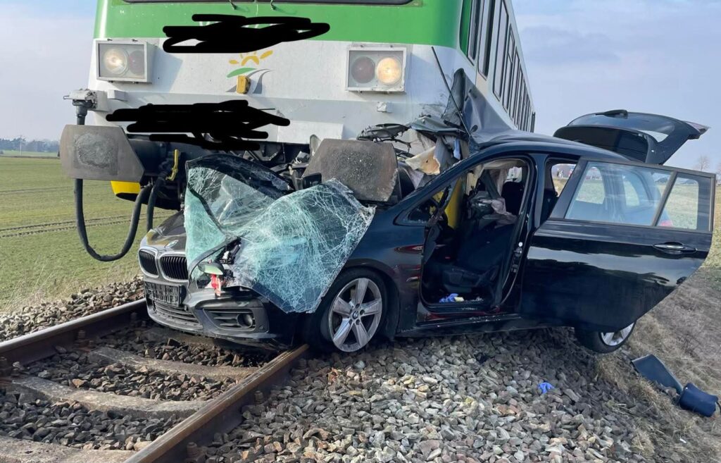 Czarny samochód osobowy stoi na torach kolejowych, zniszczony przez nadjeżdżający pociąg, z rozbitym przednim szybą.