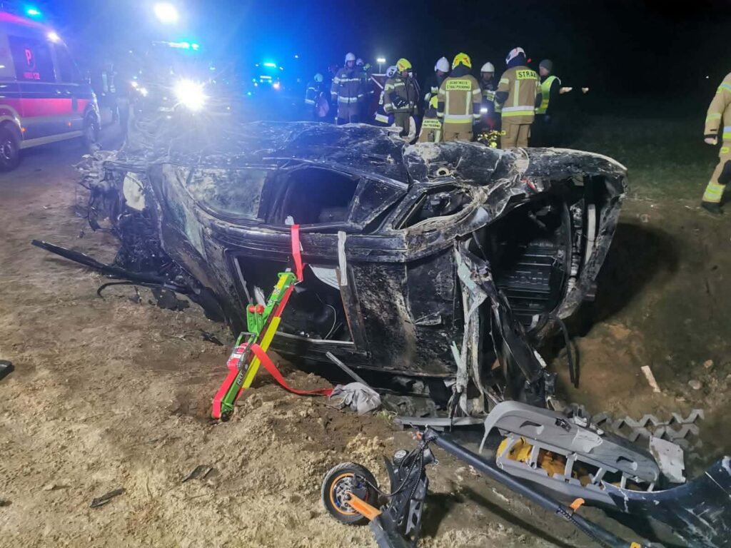 W nocy strażacy pracują przy przewróconym samochodzie osobowym na poboczu drogi, otoczonym przez radiowozy.