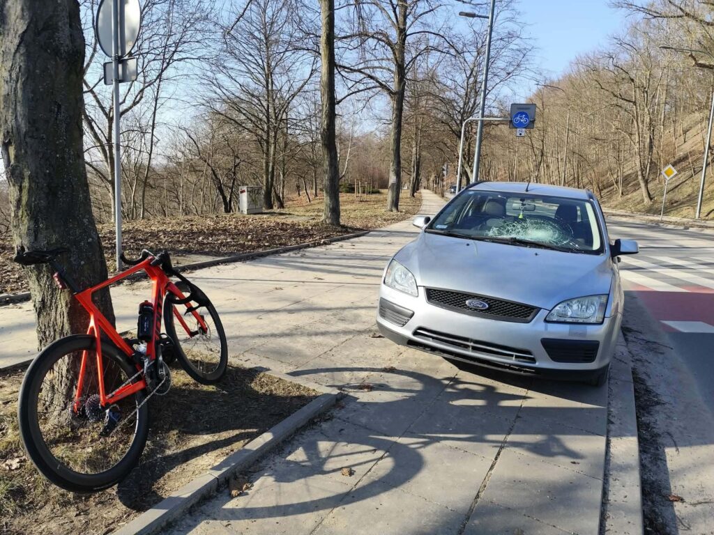 Rower w jaskrawym kolorze oparty o drzewo obok uszkodzonego samochodu zaparkowanego na chodniku w parku.