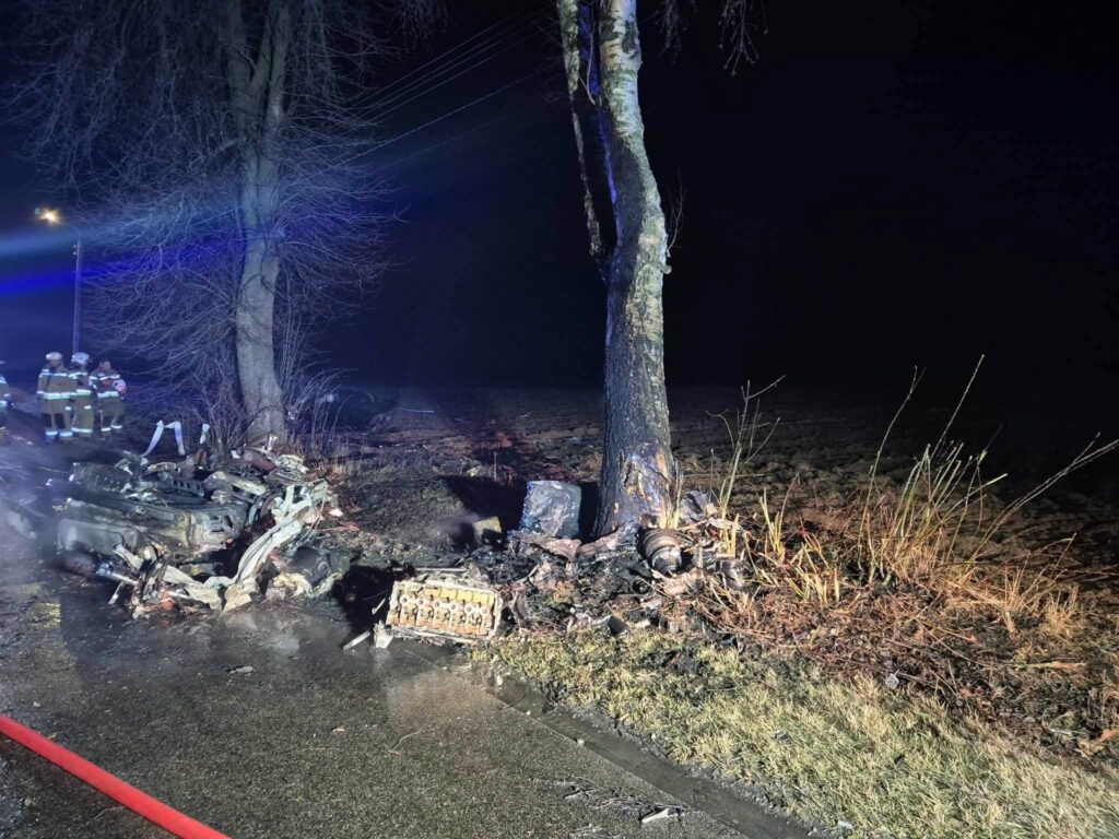 Zniszczony wrak pojazdu leży obok drzewa, a w tle widać strażaków przy pracy w ciemnej scenerii nocnej.