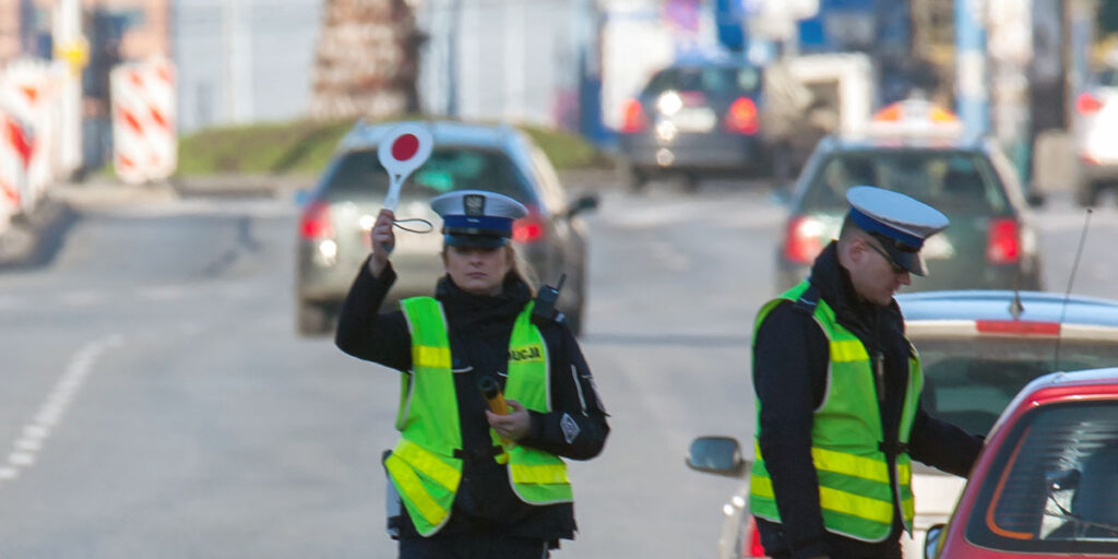 Policjantka w żółtej kamizelce kieruje ruchem na ulicy, podczas gdy drugi funkcjonariusz sprawdza dane kierowcy.