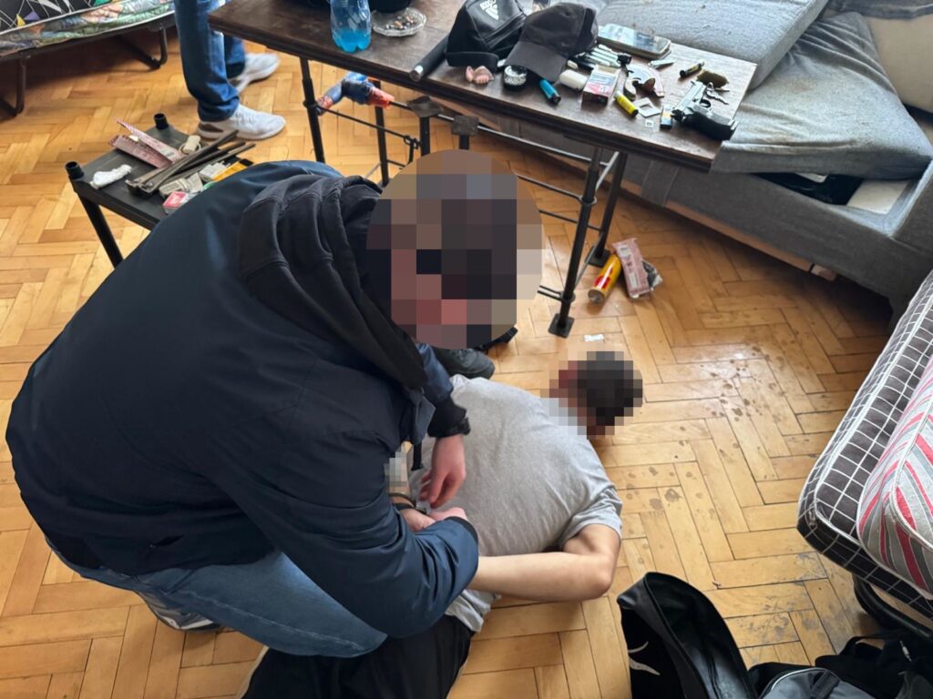 Funkcjonariusz w mundurze zatrzymuje mężczyznę leżącego na podłodze w nieuporządkowanym pomieszczeniu.