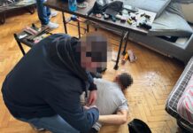 Funkcjonariusz w mundurze zatrzymuje mężczyznę leżącego na podłodze w nieuporządkowanym pomieszczeniu.