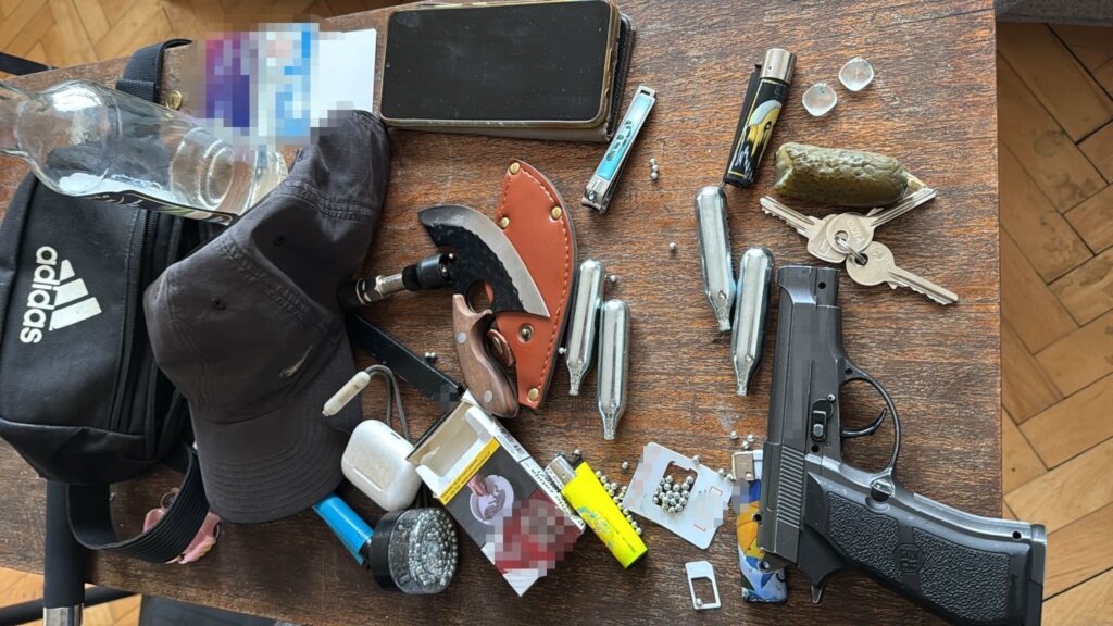 Na drewnianym stole leży różnorodne przedmioty, w tym pistolet, nóż, butelka, klucze oraz kilka zapalniczek.