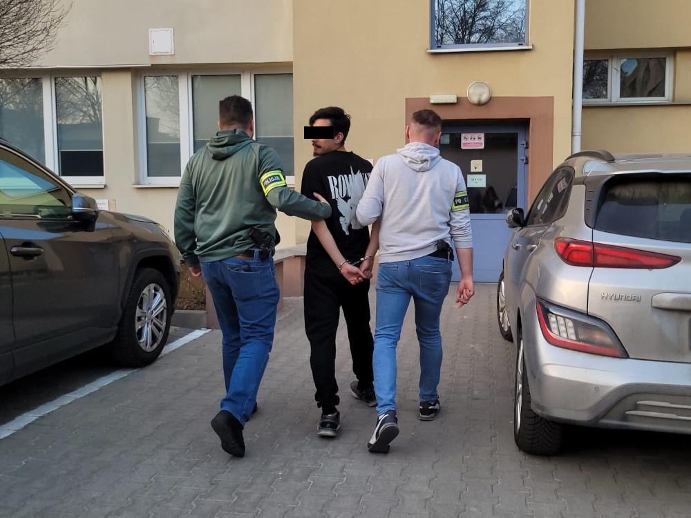 Mężczyzna w kajdankach prowadzony przez dwóch funkcjonariuszy w pobliżu budynku mieszkalnego.