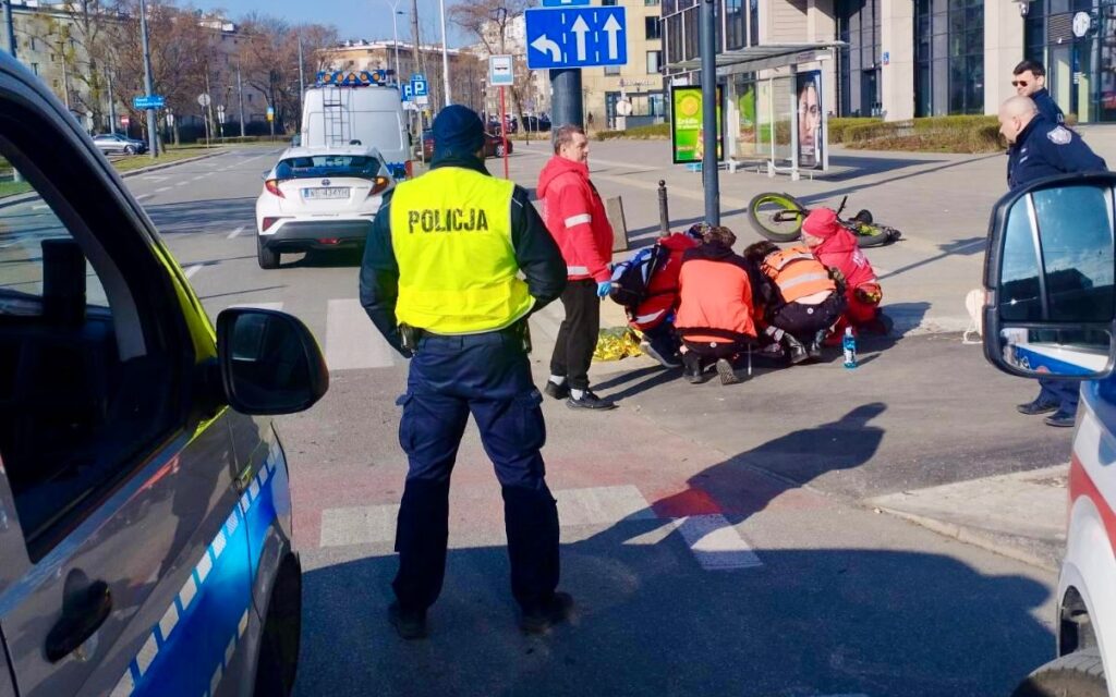 Grupa ratowników medycznych udziela pomocy poszkodowanemu na ulicy, obok stojącego radiowozu policyjnego.