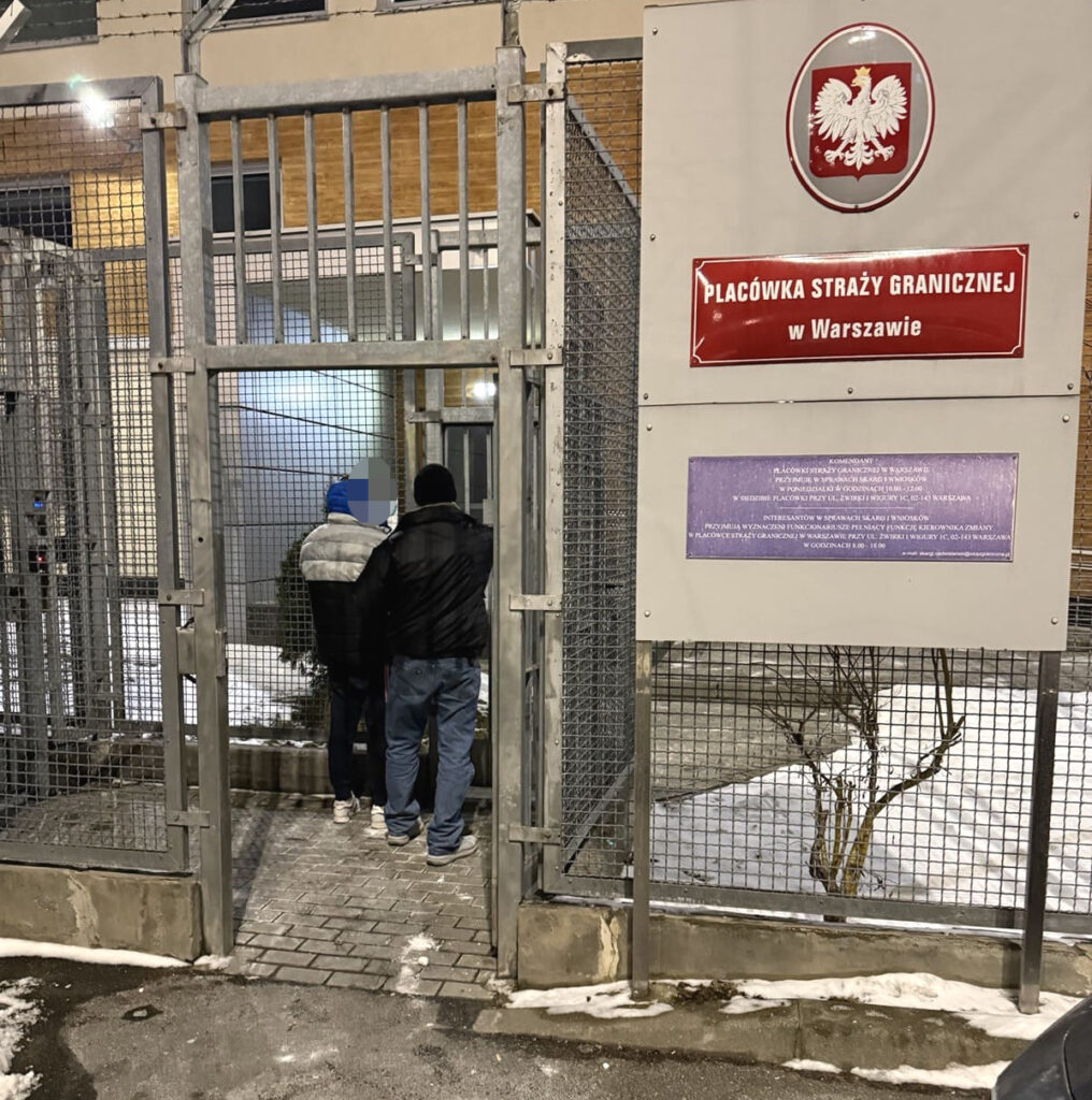 Dwaj mężczyźni stoją przy otwartych drzwiach placówki straży granicznej, w zimowym otoczeniu.