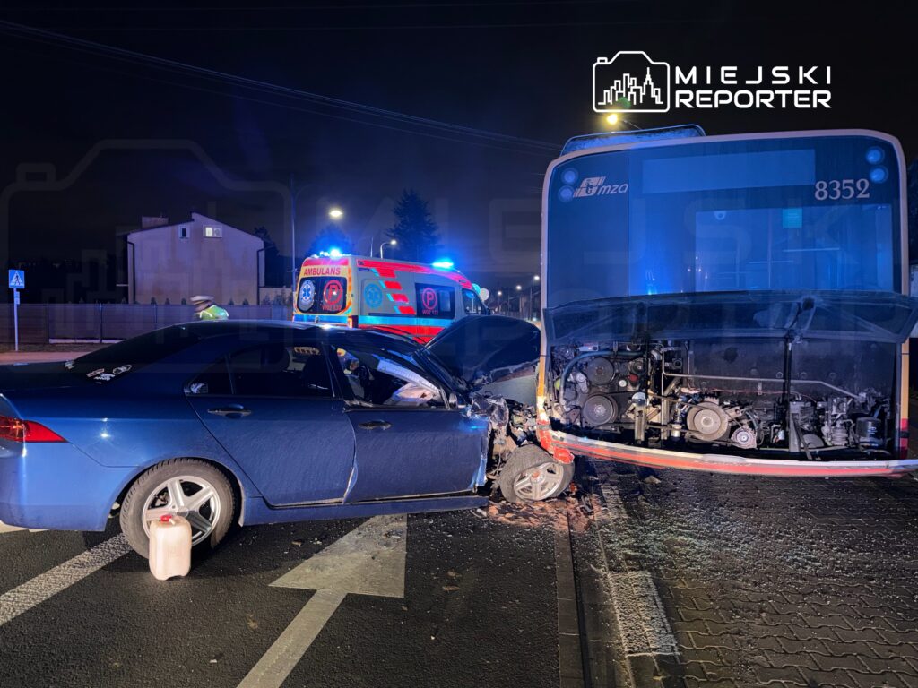 Niebieski samochód osobowy z uszkodzoną stroną zderza się z tyłem autobusu, obok stoi karetka z włączonymi światłami.