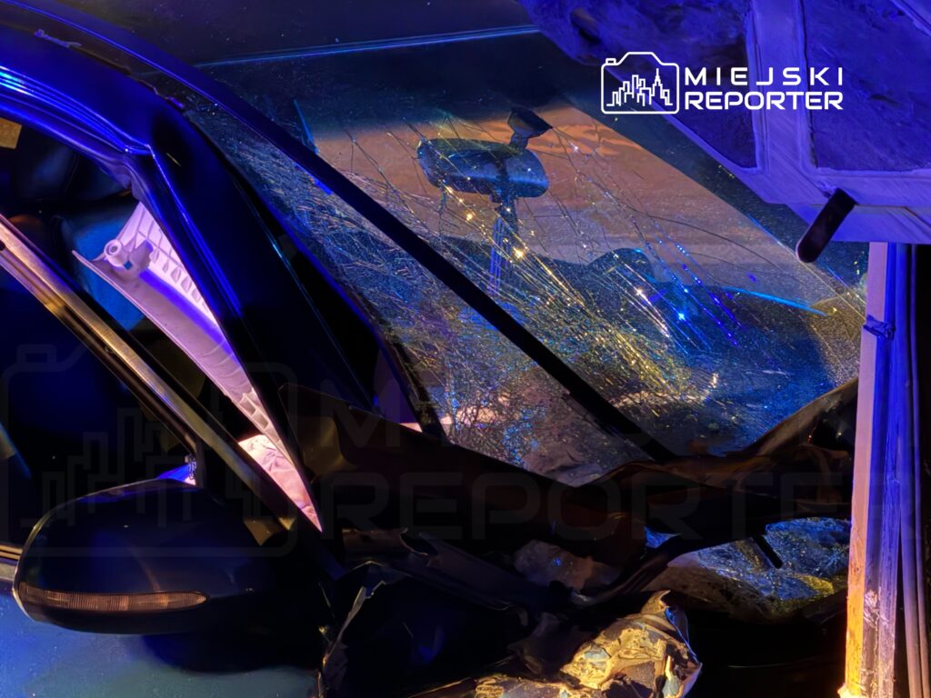 Uszkodzone auto z rozbitym oknem, znajdujące się pod niebieskim oświetleniem, w otoczeniu zniszczonych elementów.