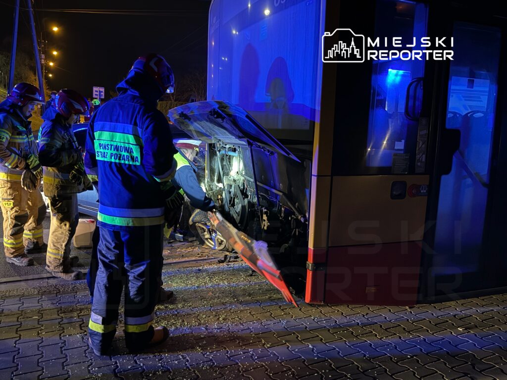 Grupa strażaków w uniformach pracuje nad usunięciem wgniecenia z samochodu osobowego po wypadku, w nocy.