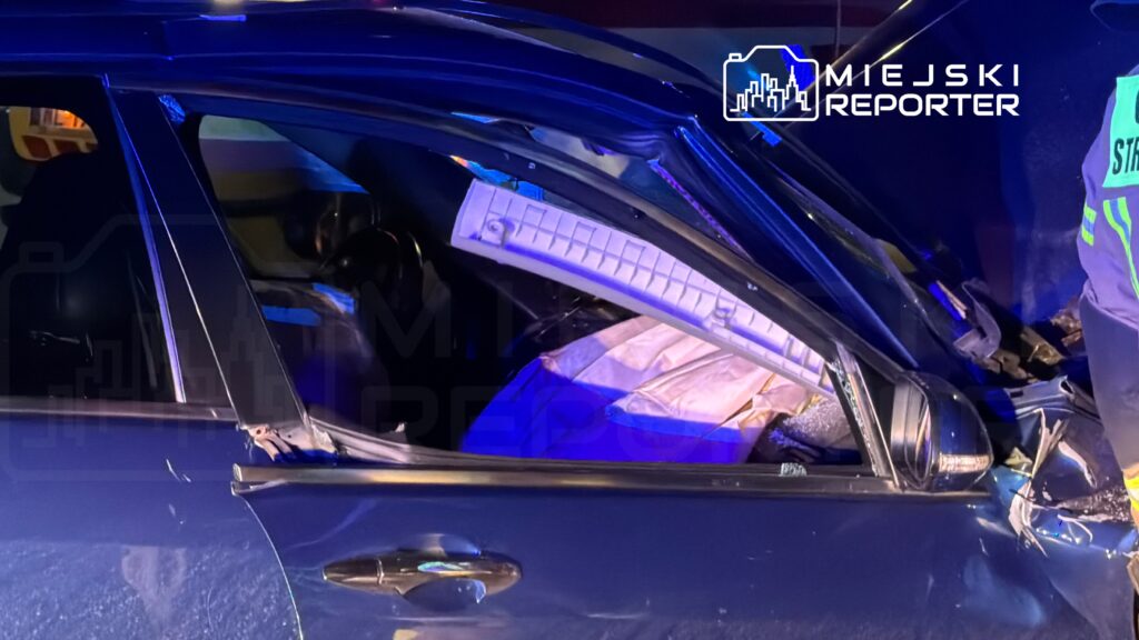 Funkcjonariusz w mundurze przeszukuje wnętrze uszkodzonego samochodu osobowego w nocy.