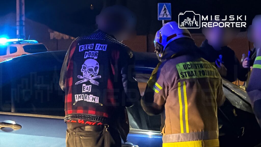 Mężczyzna w kurtce z nadrukiem "Feed the Poor, Eat the Rich" stoi obok samochodu, podczas gdy strażak udziela pomocy.