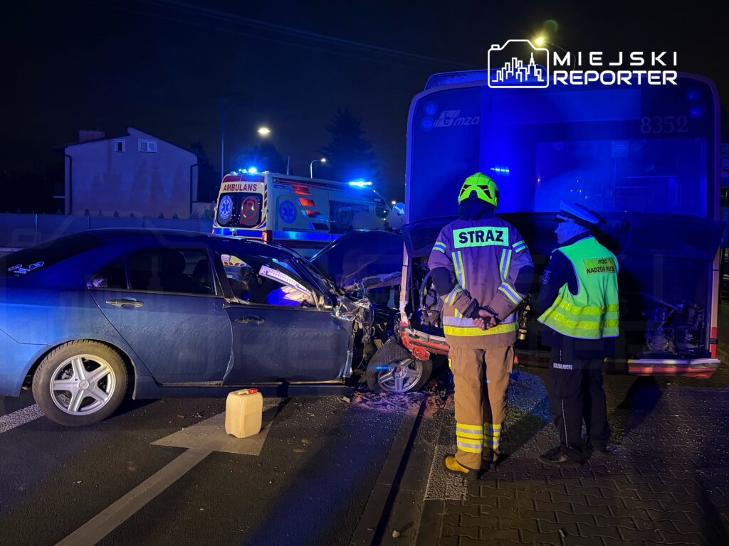 W nocy widać wypadek drogowy, w którym niebieski samochód osobowy zderzył się z autobusem. Obok stoją strażacy i policjant. W tle widoczna karetka pogotowia.