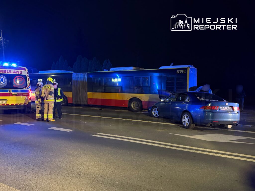 W nocy, przy drodze, strażacy rozmawiają z policjantem obok uszkodzonego autobusu i samochodu osobowego.