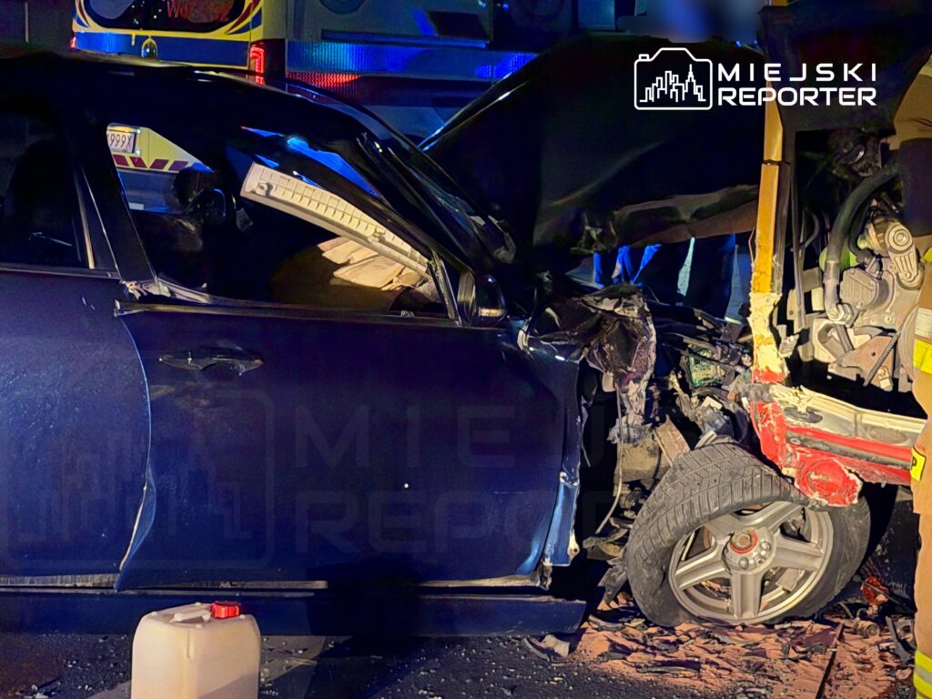 Zniszczony samochód osobowy z wgniecioną stroną, obok strażacy i służby ratunkowe na miejscu wypadku.