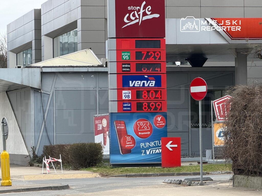 Stacja paliw z cenami paliw wyświetlanymi na dużym ekranie, w otoczeniu nowoczesnych budynków i znaków drogowych.