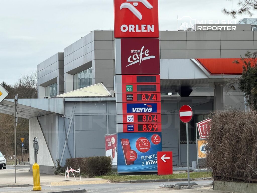 Stacja paliw z cenami paliw na wyświetlaczu, w tym 8,74 PLN za benzynę 95. W tle nowoczesny budynek.