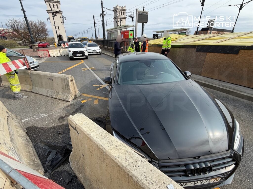 Czarny samochód osobowy stoi na drodze, obok grupy pracowników w kamizelkach odblaskowych, przy barierkach budowlanych.