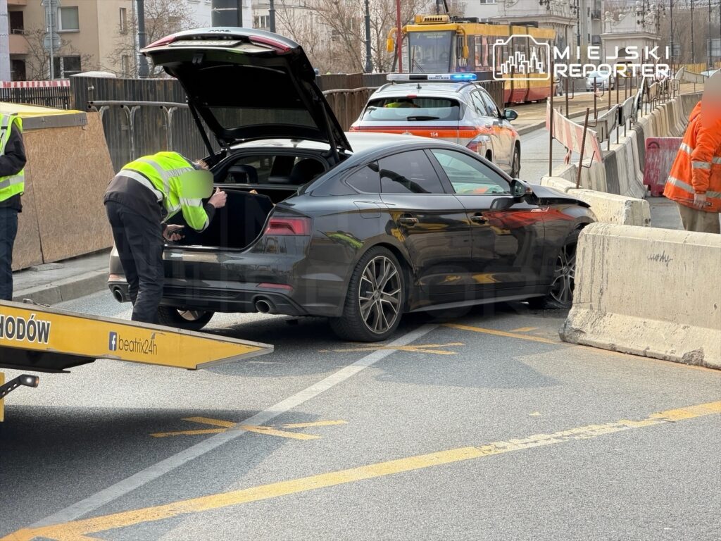Mężczyzna w odblaskowej kamizelce przeszukuje bagażnik czarnego samochodu osobowego obok barierek drogowych.
