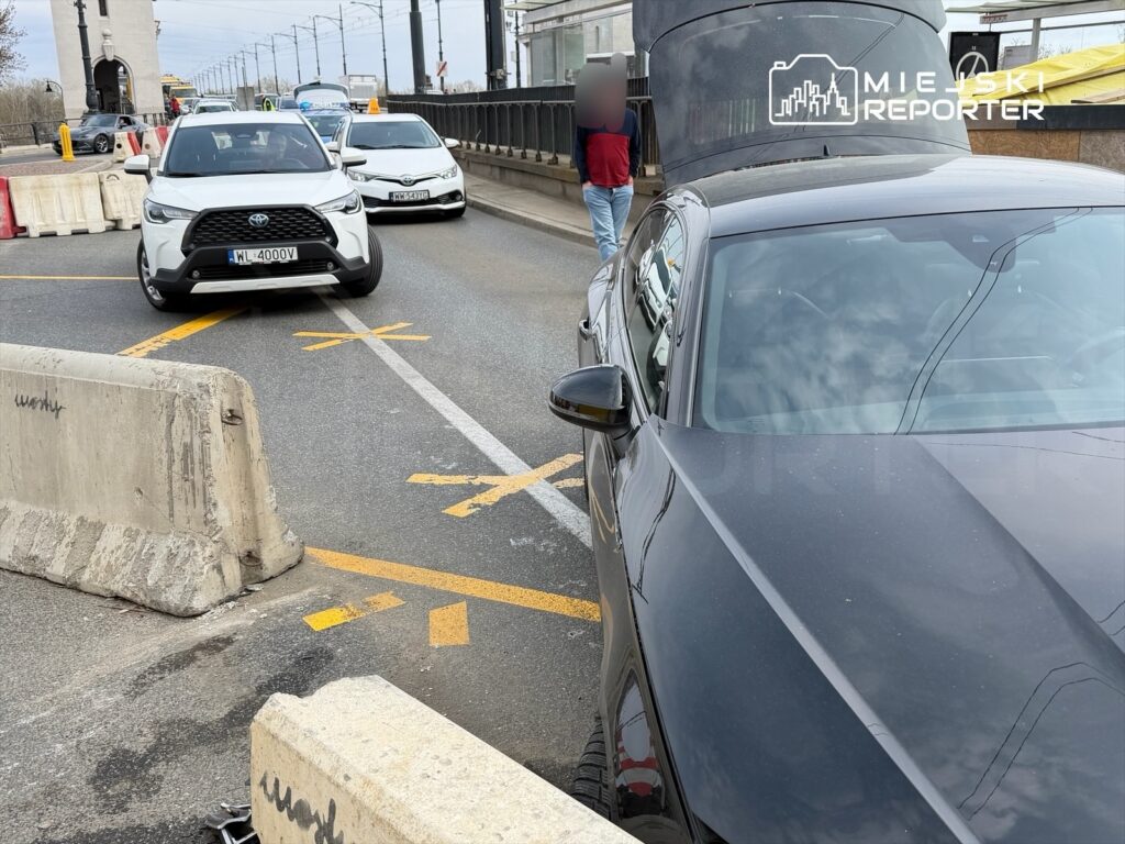 Ruch samochodowy na drodze z barierkami, z białym SUV-em skręcającym w lewo, a w tle widoczny pieszy.