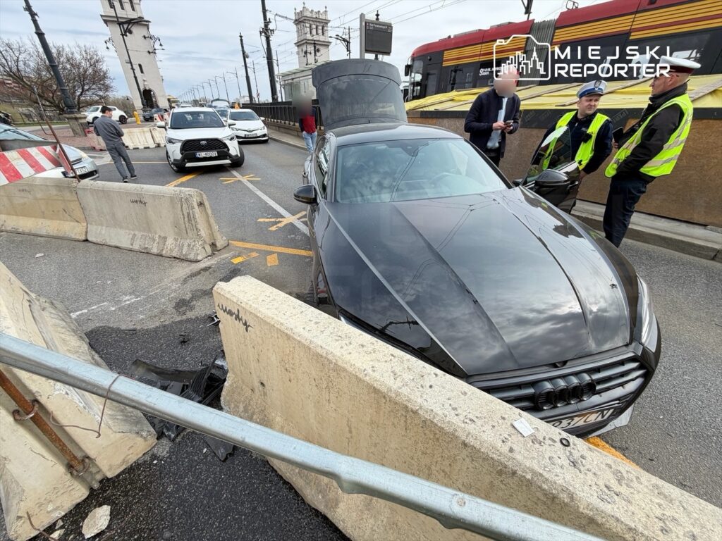 Czarny samochód osobowy stoi w zablokowanym miejscu obok betonowych blokad, a obok grupka mężczyzn w odblaskowych kamizelkach rozmawia.