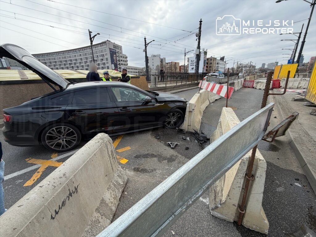 Czarny samochód osobowy stoi na drodze z otwartą klapą, obok grupka osób w odblaskowych kamizelkach rozmawia na tle barier budowlanych.
