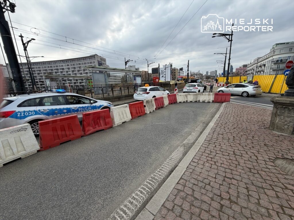 Policyjny radiowóz z niebieskimi światłami stoi na drodze, zabezpieczonej barierkami, w tle budynki miejskie.