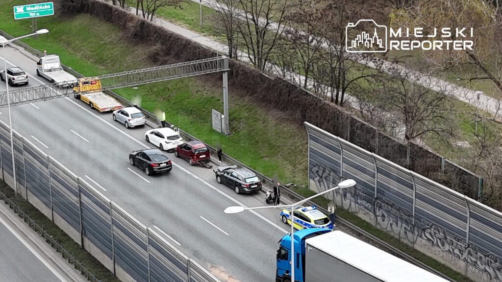 Ruch uliczny z kilkoma samochodami zaparkowanymi na poboczu, w tym policyjnym radiowozem i lawetą.