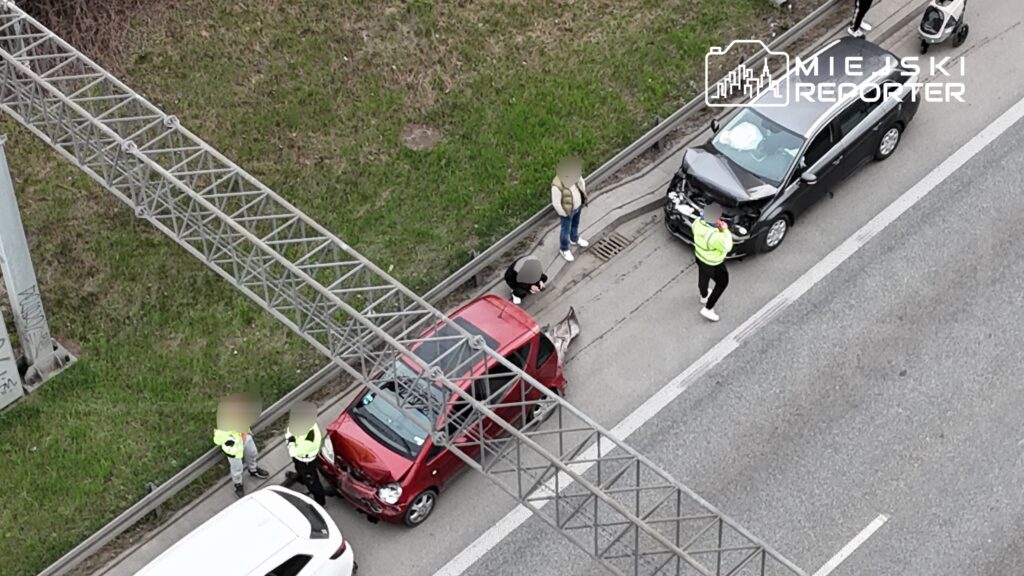 Na drodze doszło do wypadku, gdzie uszkodzone czerwone i czarne samochody stoją obok siebie, a policjanci prowadzą czynności.