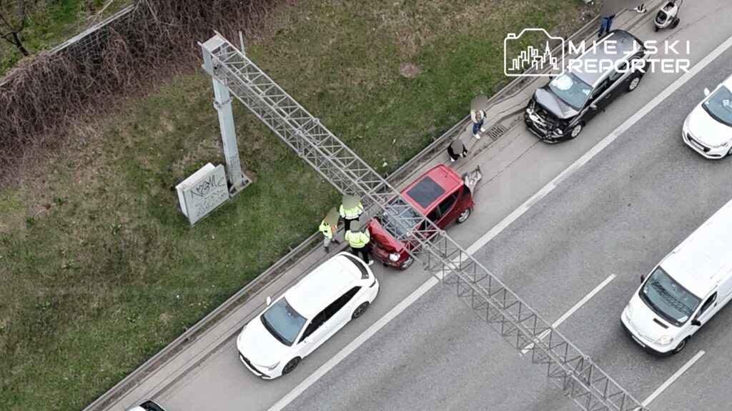Trzech pracowników w odblaskowych kamizelkach udziela pomocy przy uszkodzonym czerwonym samochodzie na poboczu drogi.