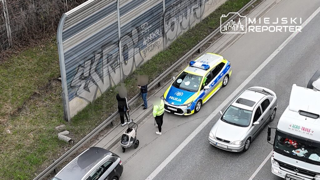 Policjant rozmawia z dwiema osobami stojącymi przy drodze obok zaparkowanego radiowozu, w pobliżu torów.