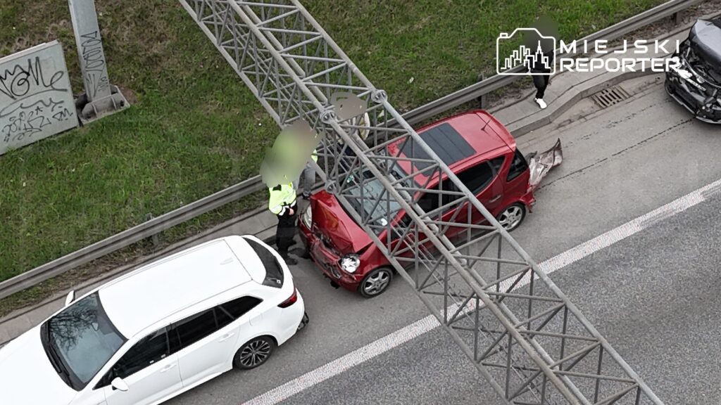 Policjant rozmawia z kierowcą przy uszkodzonym czerwonym samochodzie na drodze, obok białego pojazdu.