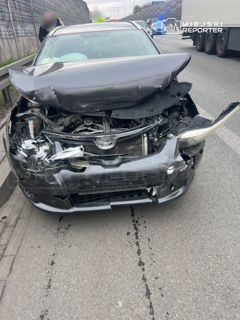 Uszkodzony samochód osobowy z wgniecionym przodem stoi na jezdni, obok widać służby ratunkowe.