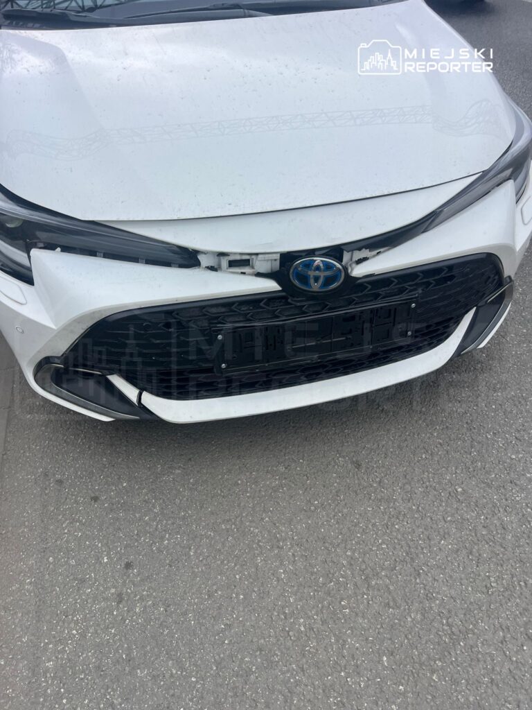 Uszkodzony biały samochód marki Toyota z uszkodzoną przednią częścią, stojący na szarej nawierzchni.